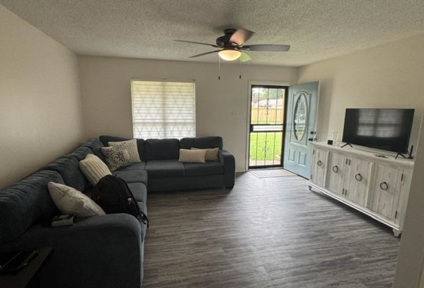 Open-concept living space with wood-finish flooring, a contemporary ceiling fan with light fixture, and a light blue front door with an oval glass insert and security screen
