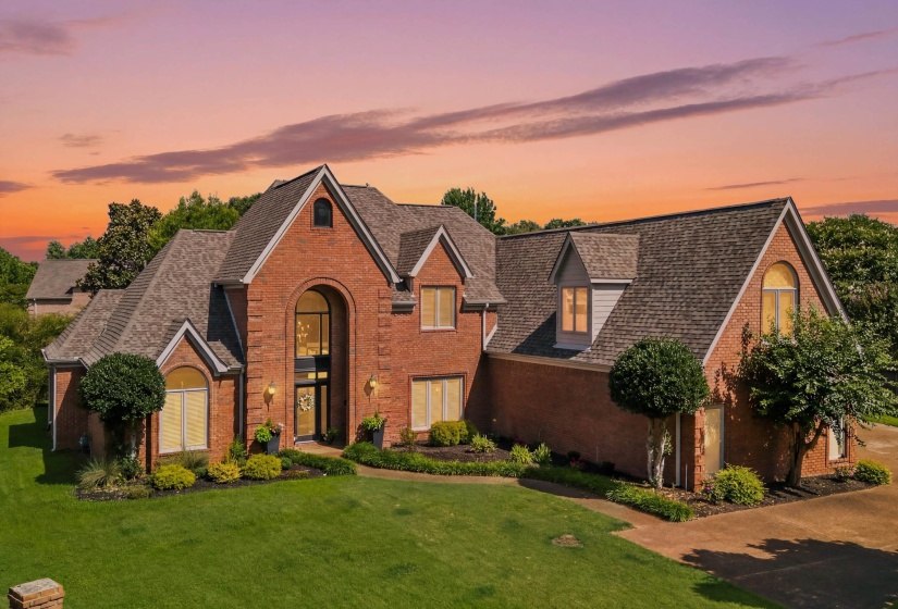 Brick exterior featuring a grand arched entry with a two-story window system, multiple gables, dormer windows, and a manicured lawn
