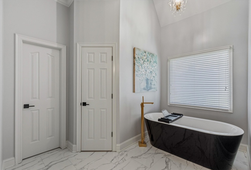 Freestanding black soaking tub with a gold-tone floor-mounted faucet, set against a window with white blinds