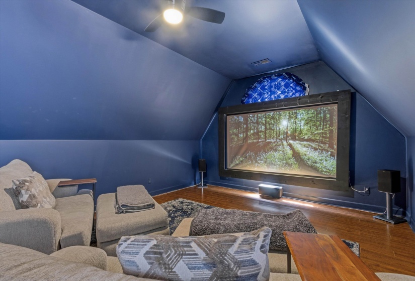 Dedicated media room featuring a large projection screen framed in dark wood, wood-finish flooring, a ceiling fan with integrated lighting, and deep blue wall finishes