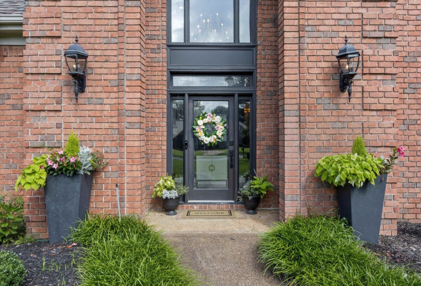 Brick exterior featuring a black framed glass entry door with transom and sidelights, flanked by two black lantern-style wall sconces and decorative planters