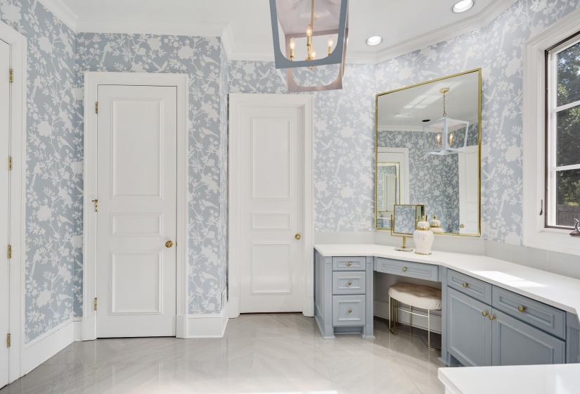 Spacious vanity area featuring light blue floral wallpaper, white countertops, built-in cabinetry with brass hardware, a large framed mirror, and recessed lighting