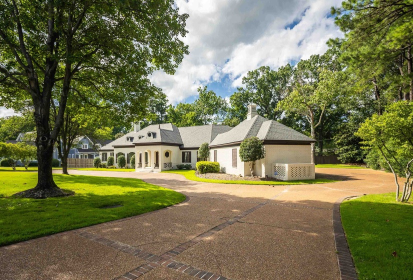 Stately residence featuring a sweeping circular driveway with brick accents, lush green lawn, mature trees, and a covered entry with columns