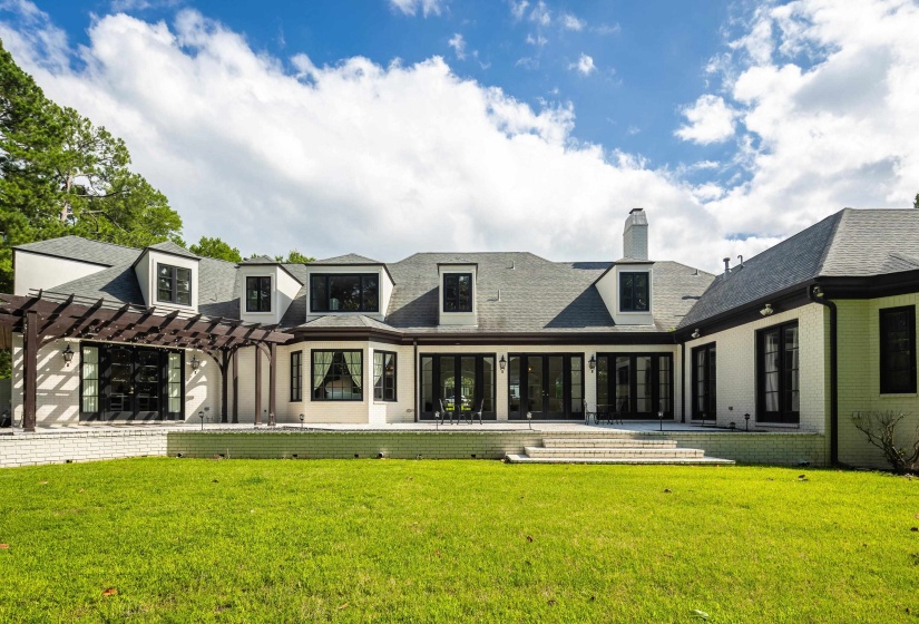Spacious backyard with a vibrant green lawn, extensive patio with multiple access points, dark wood pergola, dormer windows, and a classic brick and stucco exterior