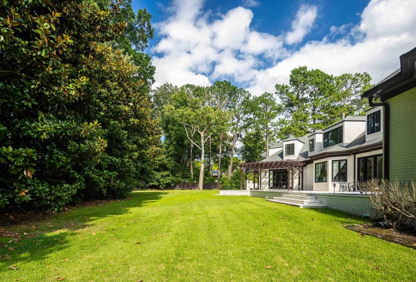 Expansive rear lawn framed by mature trees, featuring a multi-level patio with steps, an attached pergola, and multiple large windows on the home's facade