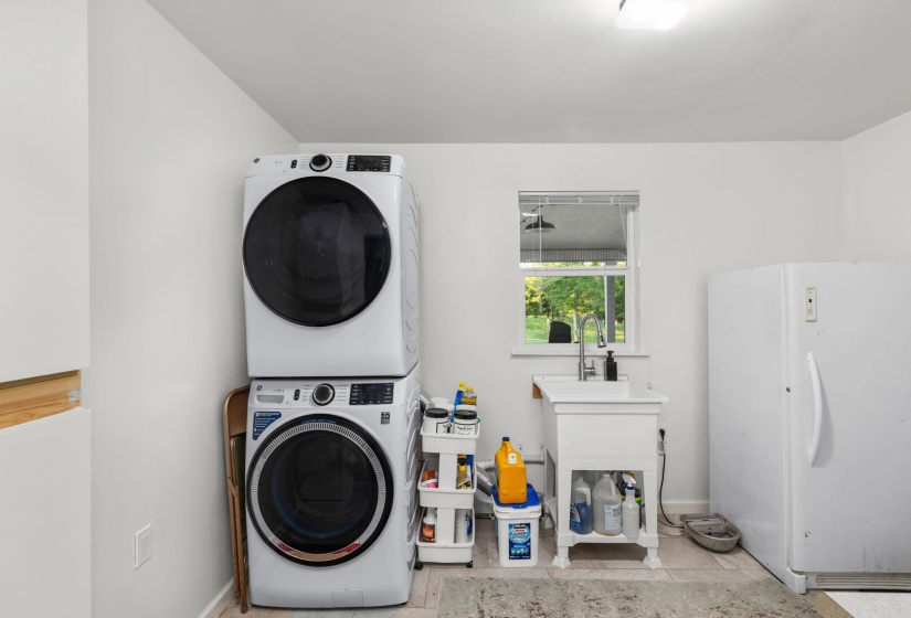 Bright utility space featuring a stacked washer and dryer, a window with natural light, a white utility sink with a chrome faucet, and a large white freestanding refrigerator