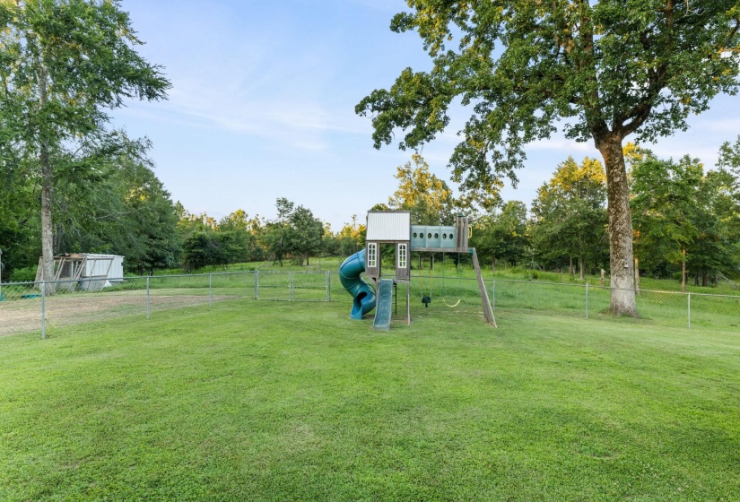 Expansive green lawn with chain link perimeter fencing, featuring mature trees and a built-in play structure with slides and swings