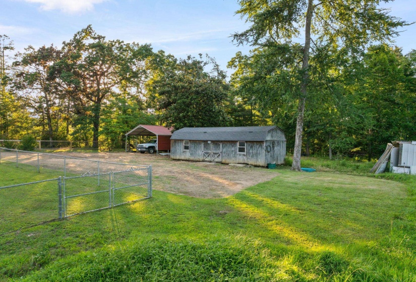 Spacious outdoor area featuring a wood-sided outbuilding with a shingled roof, a red metal carport, and a chain-link fence enclosing a portion of the property