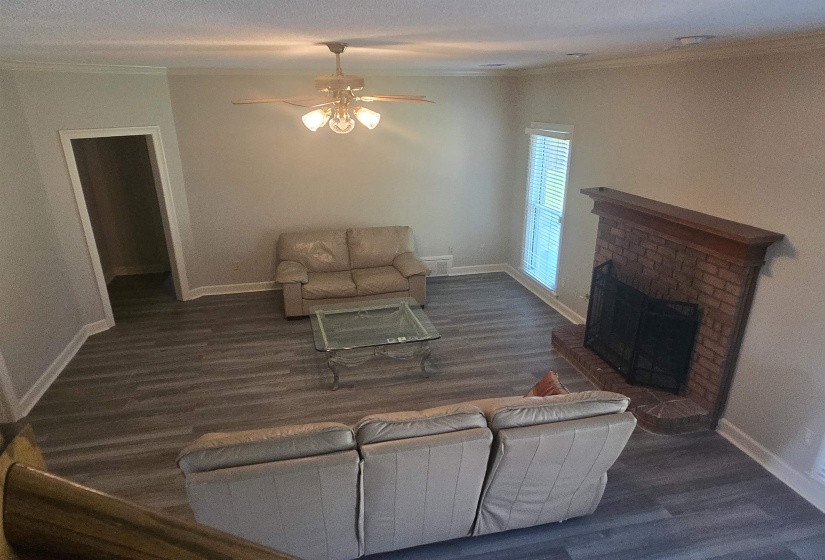 Spacious living area featuring wood-finish flooring, a brick fireplace with a mantel, a ceiling fan with integrated lighting, and crown molding