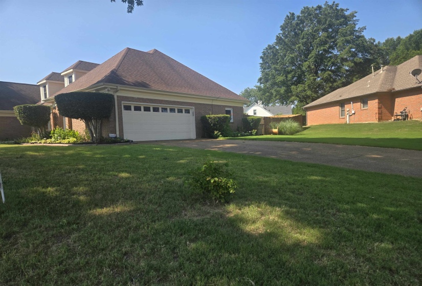 Brick exterior home featuring a two-car garage with panel detailing, a brown shingle roof, and mature perimeter landscaping