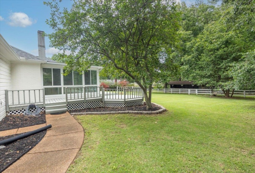 Expansive lawn area featuring a white picket fence, mature trees, and a concrete pathway leading to a screened-in porch with an attached wooden deck
