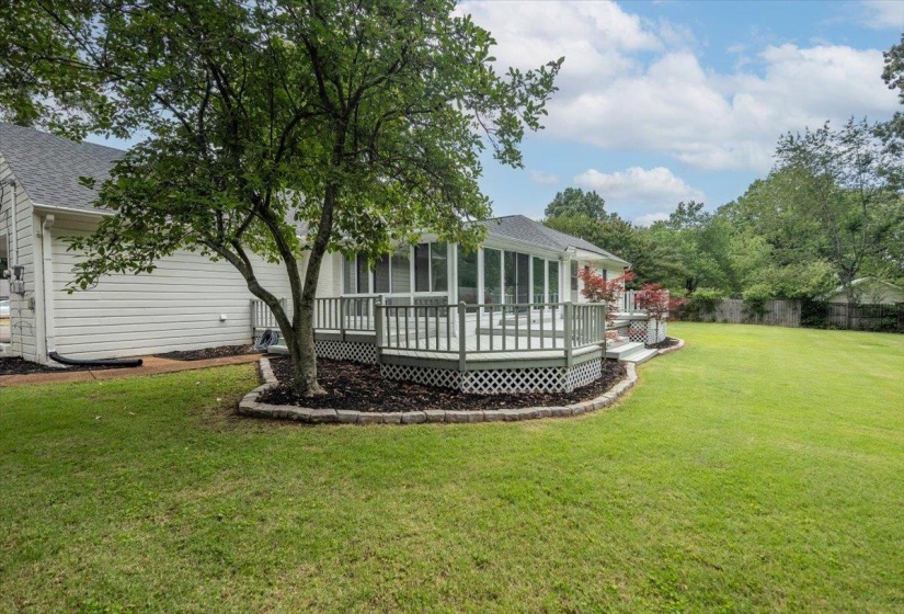 Expansive green lawn featuring a multi-level deck with white railings and lattice skirting, a screened porch, mature tree, and landscaped planting beds