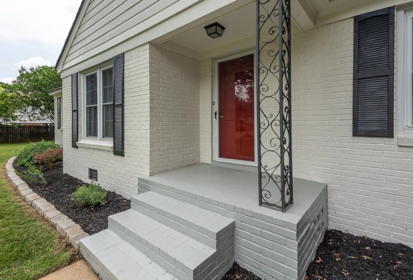 Welcoming entryway featuring a red front door, painted brick facade, decorative wrought iron column, and a neatly mulched landscape bed with low-maintenance plantings