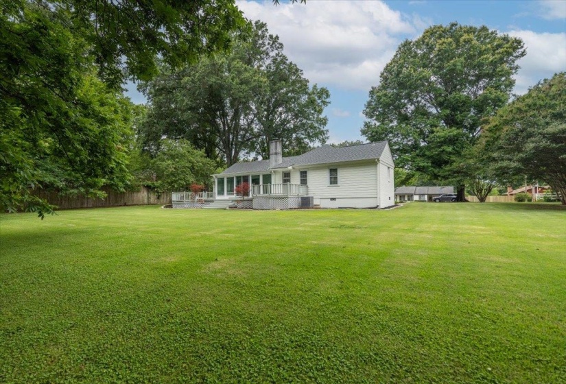 Expansive green lawn surrounds a white siding residence featuring a sunroom, a multi-level patio, and mature perimeter trees