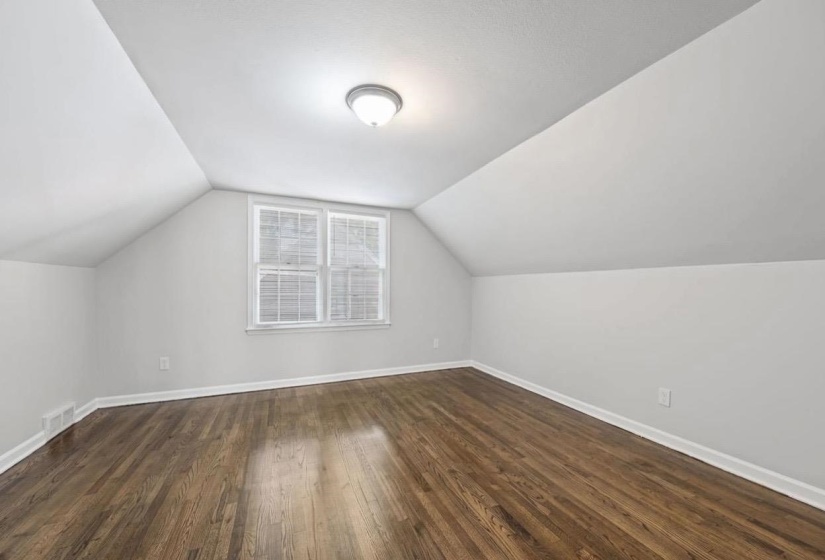 Attic room featuring sloped ceilings, a double window with blinds, wood-finish flooring, white baseboards, and a flush-mount ceiling light fixture