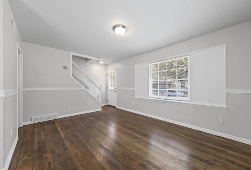 Spacious room featuring rich wood-finish flooring, a prominent window with grid detailing, and a staircase with white railings