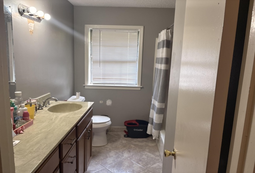 Bathroom featuring a single vanity with wood-finish cabinetry and a light-toned countertop, a window with blinds, a toilet, a built-in bathtub with shower, and tiled flooring