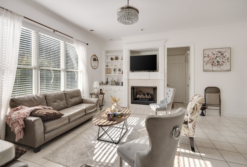Living area featuring large windows with blinds and sheer curtains, a built-in fireplace with a mantel, integrated shelving, and light-toned tile flooring
