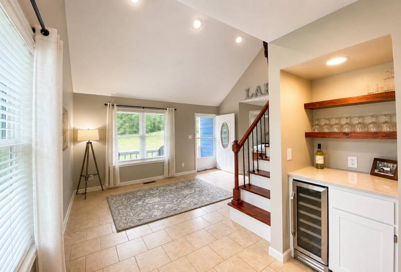 Vaulted ceiling entryway featuring large windows, neutral tile flooring, a built-in beverage bar with open shelving, and a prominent staircase with wood treads and iron balusters