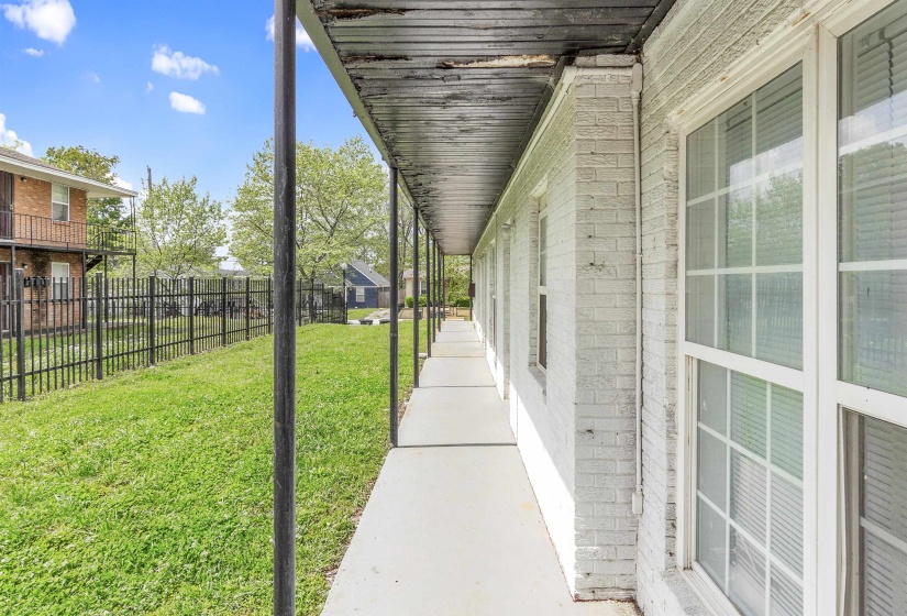 Covered walkway with painted brick exterior, multiple windows with white frames, and a perimeter fence enclosing a grassy area