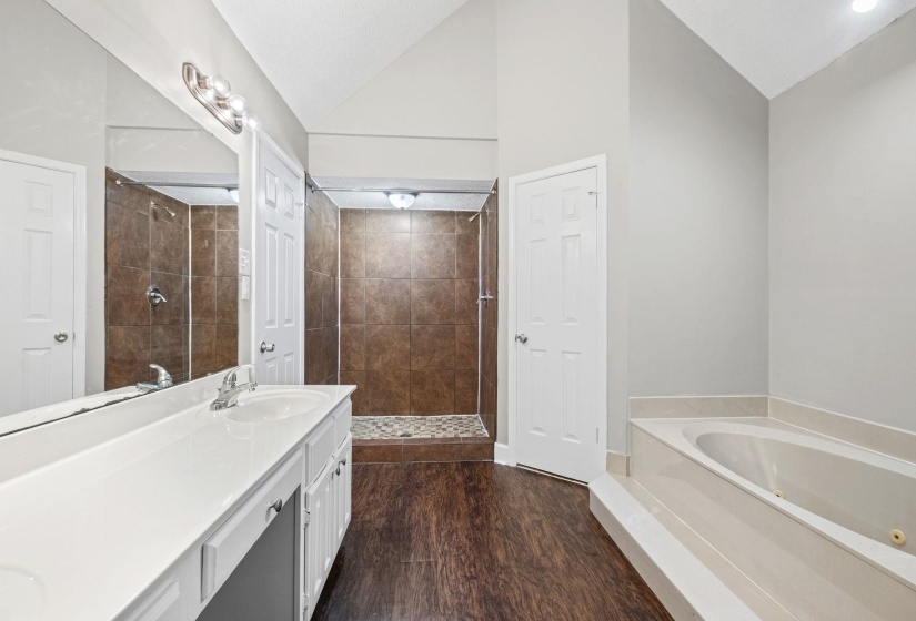 Spacious bathroom featuring a dual vanity with white cabinetry and a white countertop, a walk-in shower with brown tile surround and river rock flooring, a built-in soaking tub, and wood-finish flooring