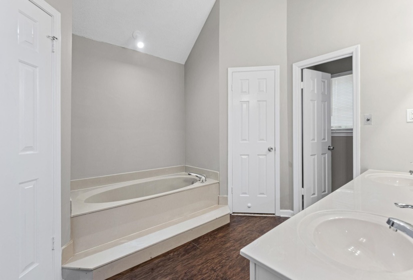 Bathroom featuring a built-in tub with a light-colored surround, vaulted ceiling with recessed lighting, wood-finish flooring, and a double vanity with undermount sinks and chrome fixtures