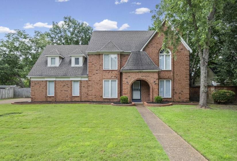 Two-story brick exterior featuring a prominent arched entry, multiple gabled dormers, a curved stone pathway, and mature landscaping