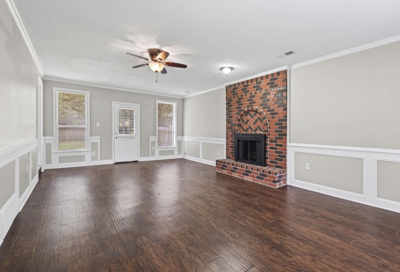 Spacious living area featuring a brick fireplace with a chevron pattern, rich wood-finish flooring, and crisp wainscoting