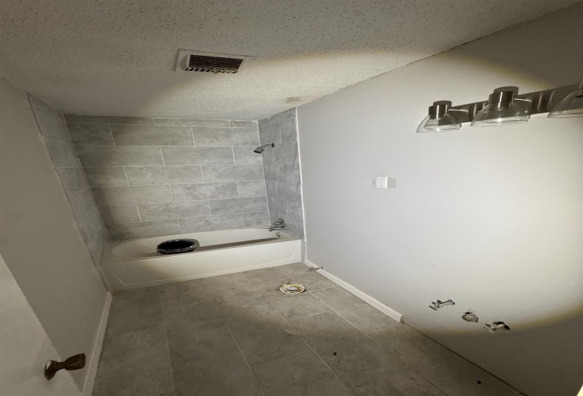 Bathroom featuring a white bathtub with a tiled shower surround, gray tile flooring, and a multi-bulb vanity light fixture