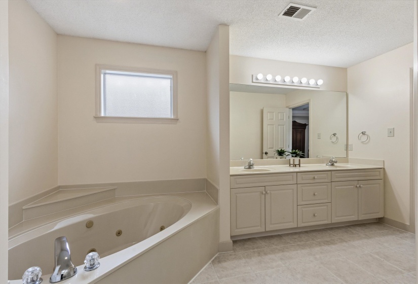 Spacious bathroom featuring a corner jetted soaking tub with steps, a large double vanity with ample storage, a wide mirror, and light-toned tile flooring