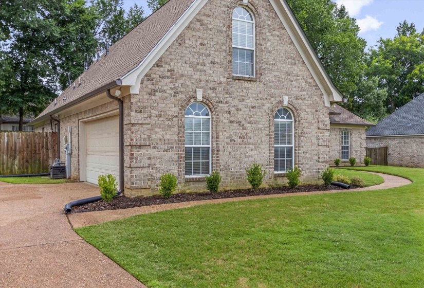 Brick exterior featuring arched windows, an attached garage, a concrete driveway, a sidewalk, and landscaping