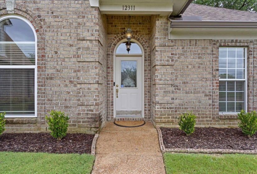 Brick facade home featuring an arched entry with a white door, an arched window, a rectangular window with grilles, and landscaping with evergreen shrubs