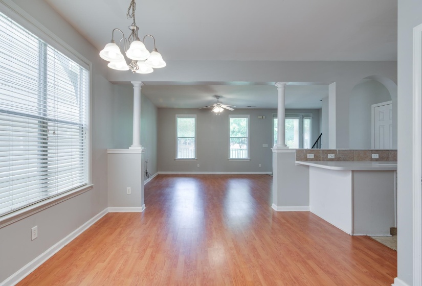 Spacious room featuring wood-finish flooring, neutral gray walls, multiple large windows with blinds, a multi-light chandelier, and decorative half-columns defining an open transition to an additional living space