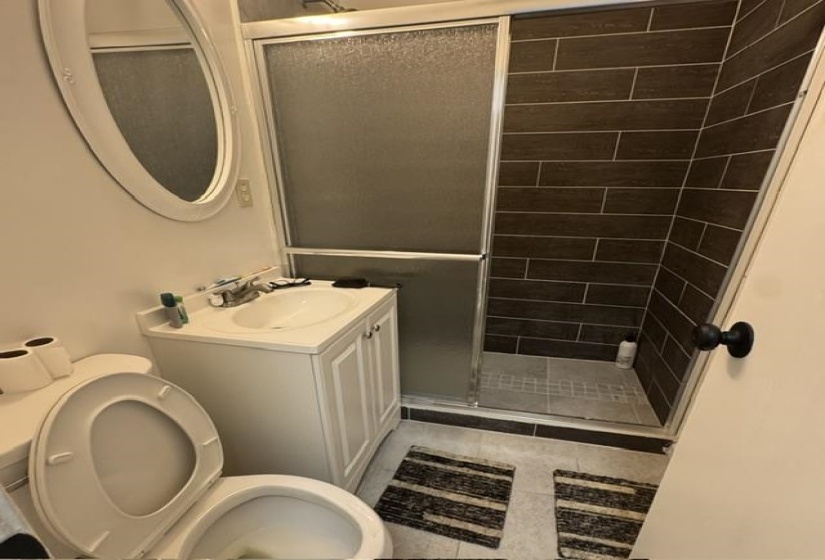 Bathroom featuring a white vanity with integrated sink, chrome faucet, oval mirror, toilet, and a stand-up shower with dark rectangular wall tiles and a glass sliding door