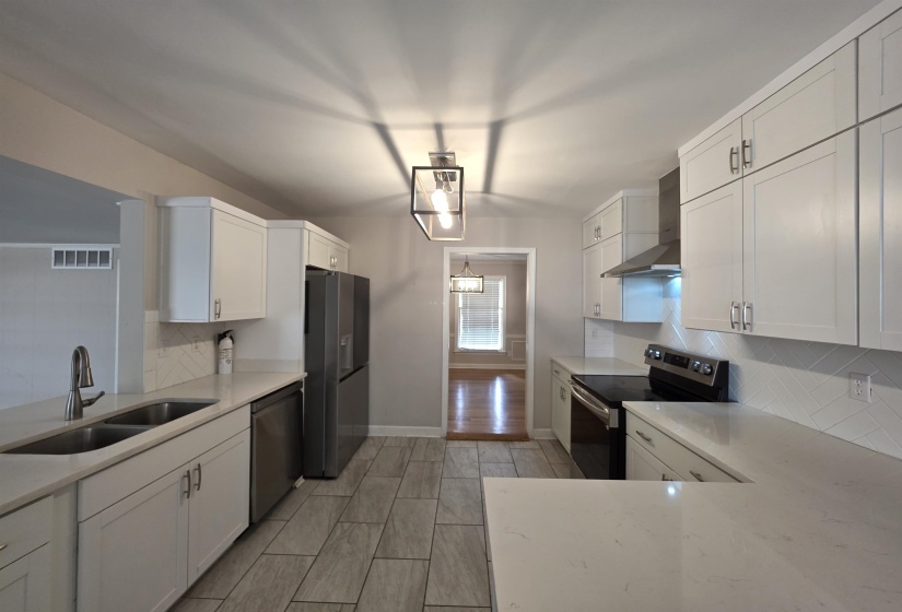 Updated kitchen featuring white shaker-style cabinetry, light-toned countertops, stainless steel appliances, a herringbone pattern backsplash, and wood-finish tile flooring
