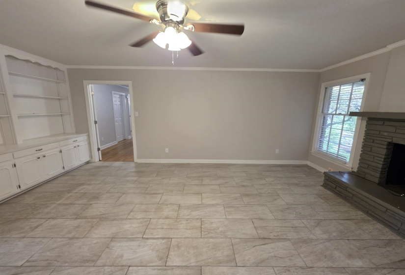 Spacious room featuring tile flooring, a built-in stone fireplace, a window with blinds, a ceiling fan with light fixture, and built-in cabinetry with open shelving