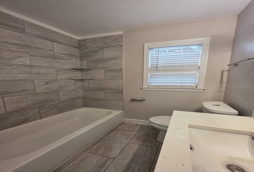 Bathroom featuring a tub with stone-look tile surround and two glass corner shelves