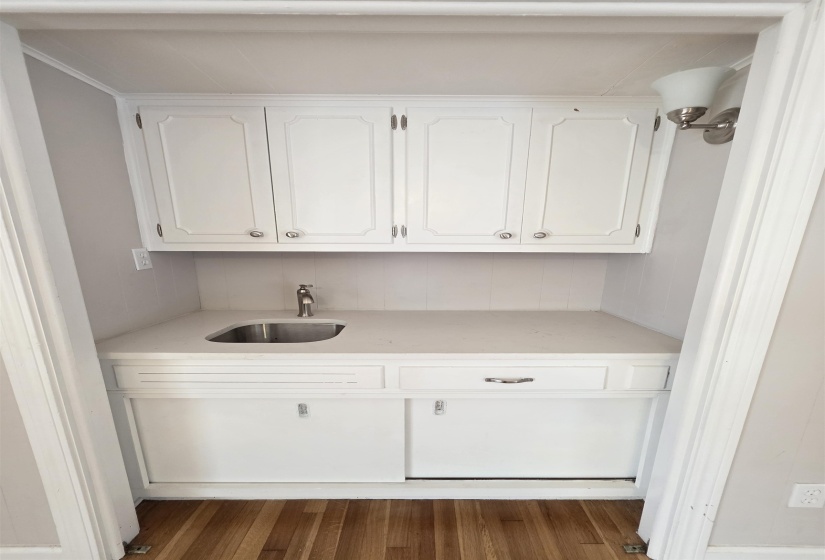 Built-in wet bar featuring white cabinetry, a stainless steel sink, a single-handle faucet, a light-colored countertop, and wood-finish flooring