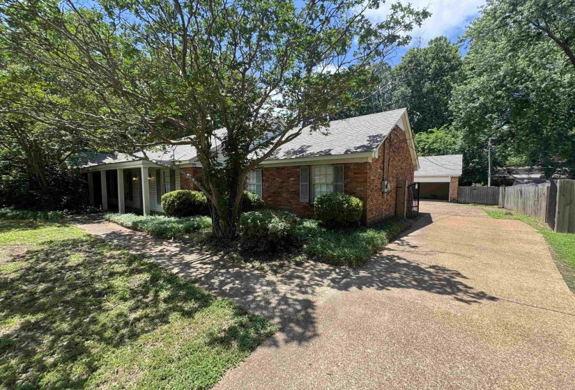Brick exterior residence featuring a covered front porch with white columns, a pitched shingle roof, mature landscaping, and a paved driveway leading to an attached garage