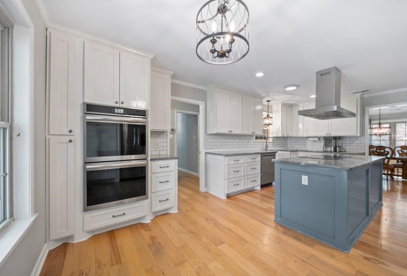 Spacious kitchen featuring white cabinetry, integrated new stainless steel double ovens, a central island with a contrasting base and granite countertop, and a stainless steel range hood