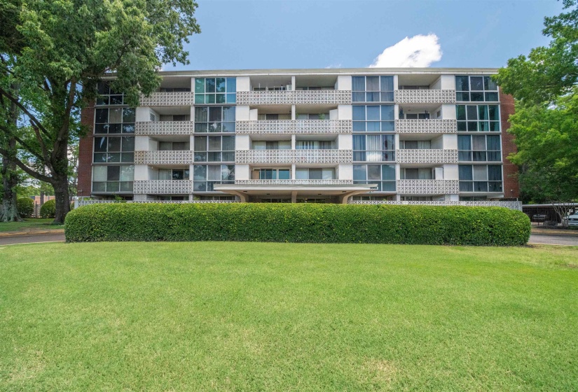 Multi-story building featuring brick and white paneling, individual balconies with decorative screens, a prominent covered entrance, and a well-maintained foreground lawn with mature hedges