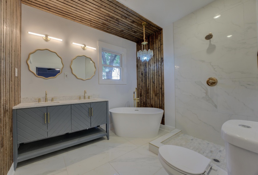 Freestanding soaking tub with a crystal chandelier, dual vanity with brass fixtures, wood-slat accent wall, walk-in shower with large format tile, and mosaic tile shower pan