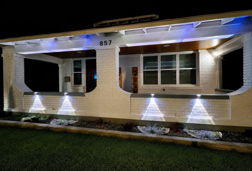 Exterior featuring white brick siding, a covered porch with decorative pillars, and accent lighting