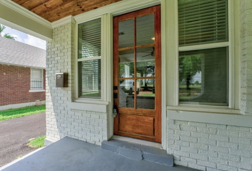 Covered porch entrance featuring a wooden front door with glass panels, white painted brick exterior, and a wood-paneled ceiling