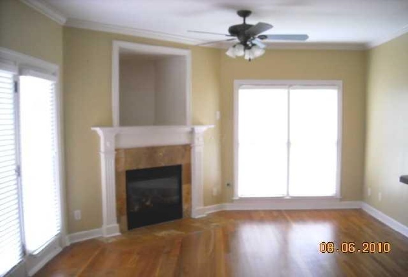 Living area featuring wood-finish flooring, a fireplace with a stone surround and white mantel, crown molding, a ceiling fan with integrated lighting, and multiple large windows