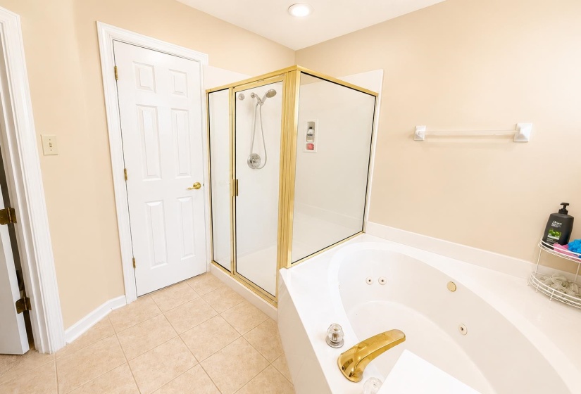 Bathroom featuring a jetted soaking tub, a separate glass-enclosed shower with gold-toned framing, light-toned wall paint, and square floor tiling