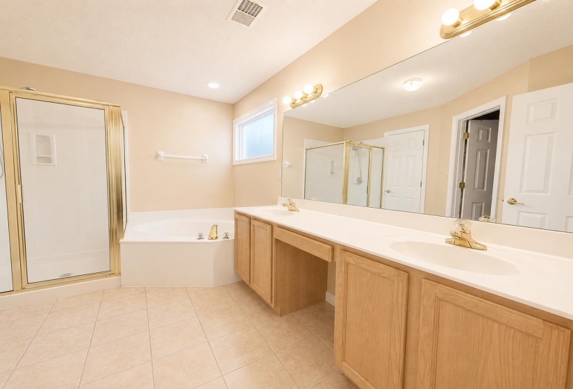 Primary Bathroom featuring a dual vanity with light wood cabinetry, a large vanity mirror, a corner soaking tub, a stand-up shower with a glass enclosure, and tiled flooring