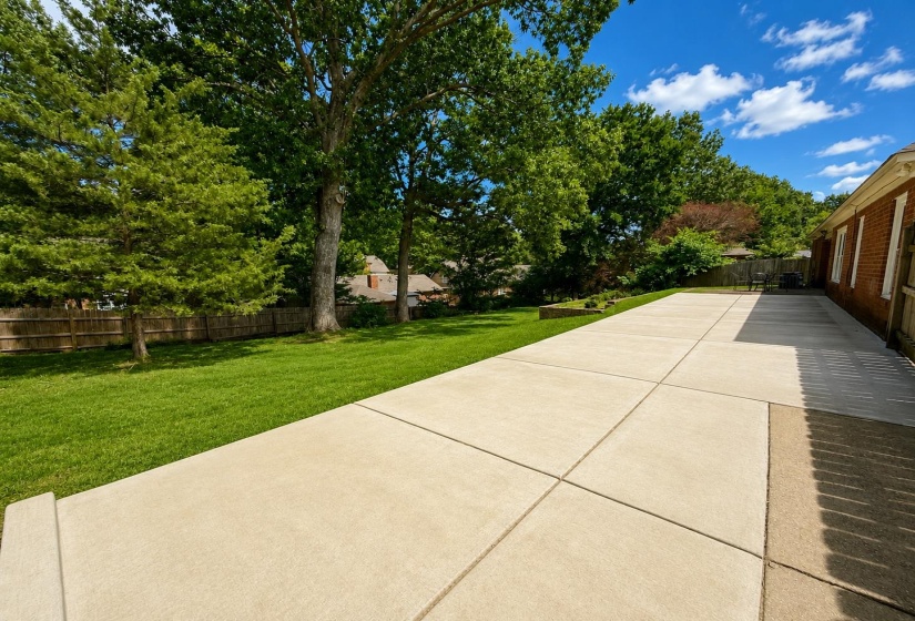 Expansive concrete patio with a manicured lawn, mature trees, and a privacy fence