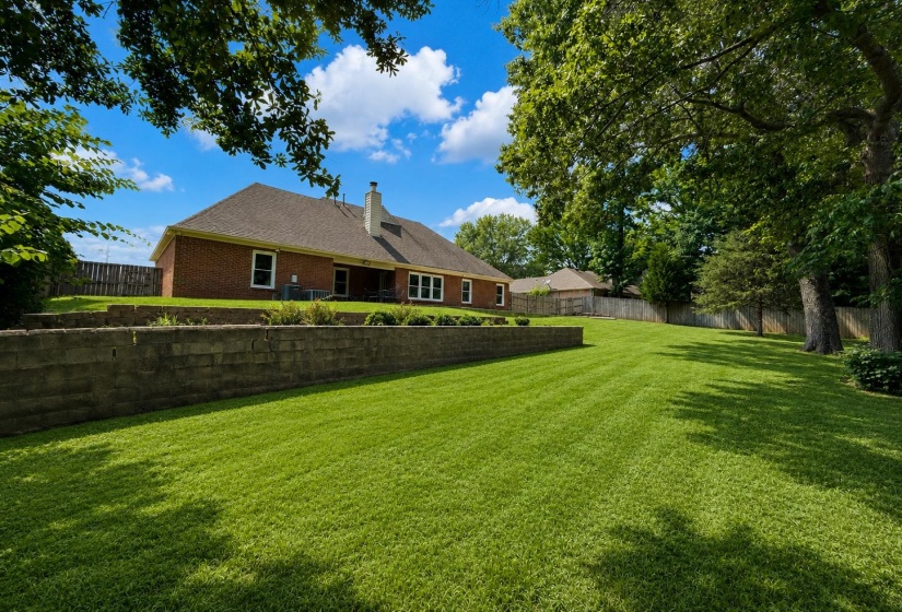 Expansive backyard featuring a lush green lawn, mature trees providing natural shade, and a brick retaining wall with landscaping