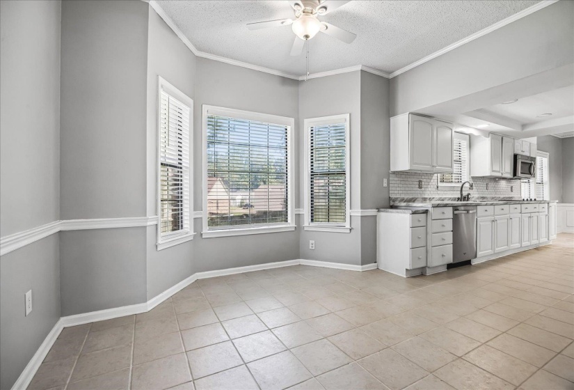 Open-concept dining area featuring tiled flooring, a bay window with blinds, and a ceiling fan with light fixture, adjacent to a kitchen with white cabinetry, stainless steel appliances, and a subway tile backsplash